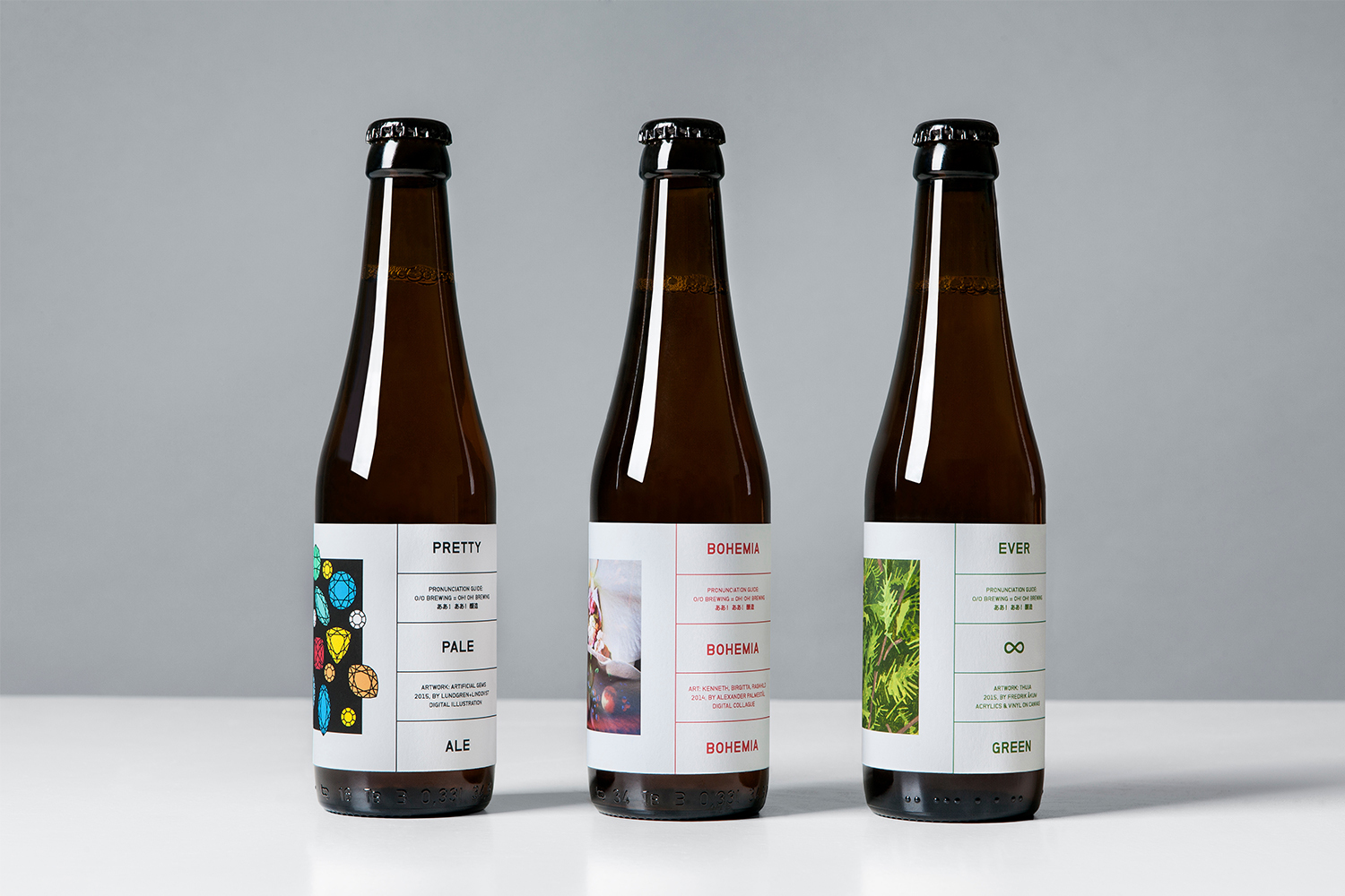 Three brown glass beer bottles with white labels featuring typography and graphic artwork standing on a white surface against a grey backdrop