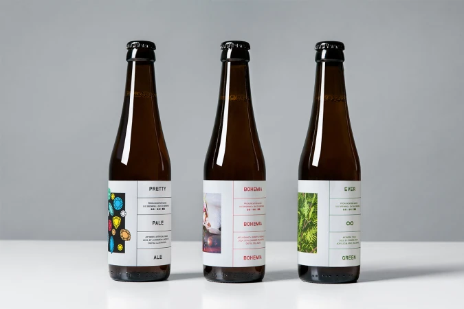 Three brown glass beer bottles with white labels featuring typography and graphic artwork standing on a white surface against a grey backdrop