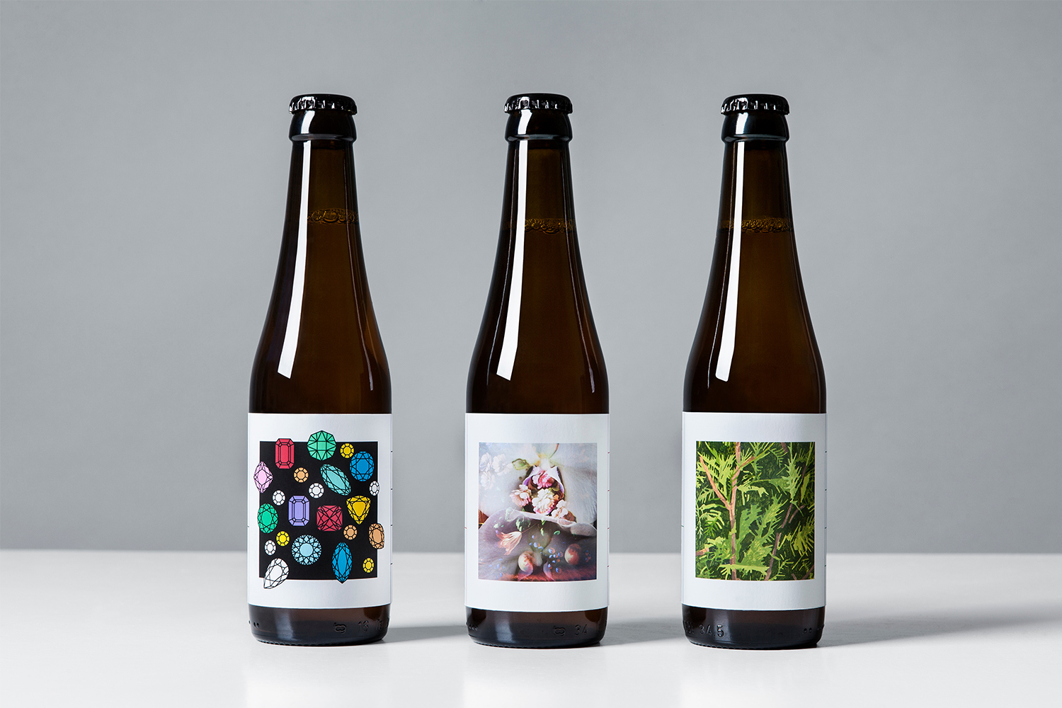 Three dark glass beer bottles with white custom art labels standing on a white surface against a light grey background.