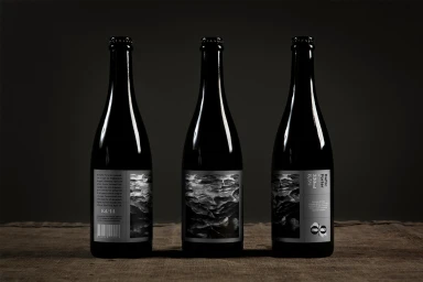 Three dark beer bottles featuring monochrome textured labels arranged upright on a dark textured fabric surface