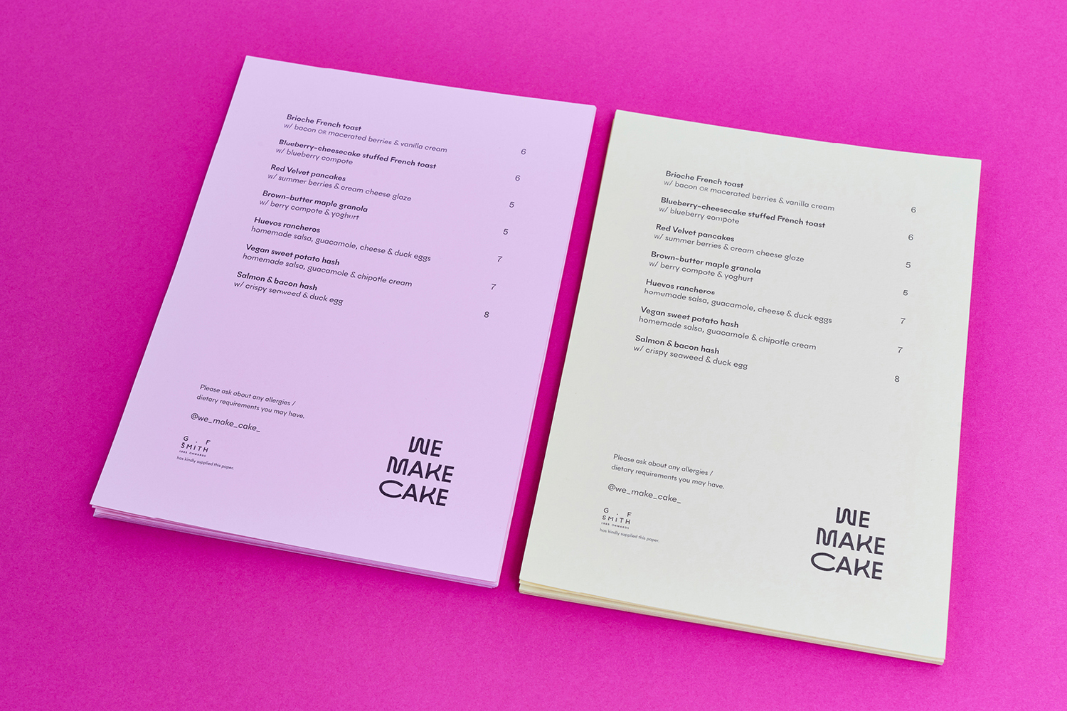 Two printed menus for We Make Cake displayed on a vibrant pink background surface