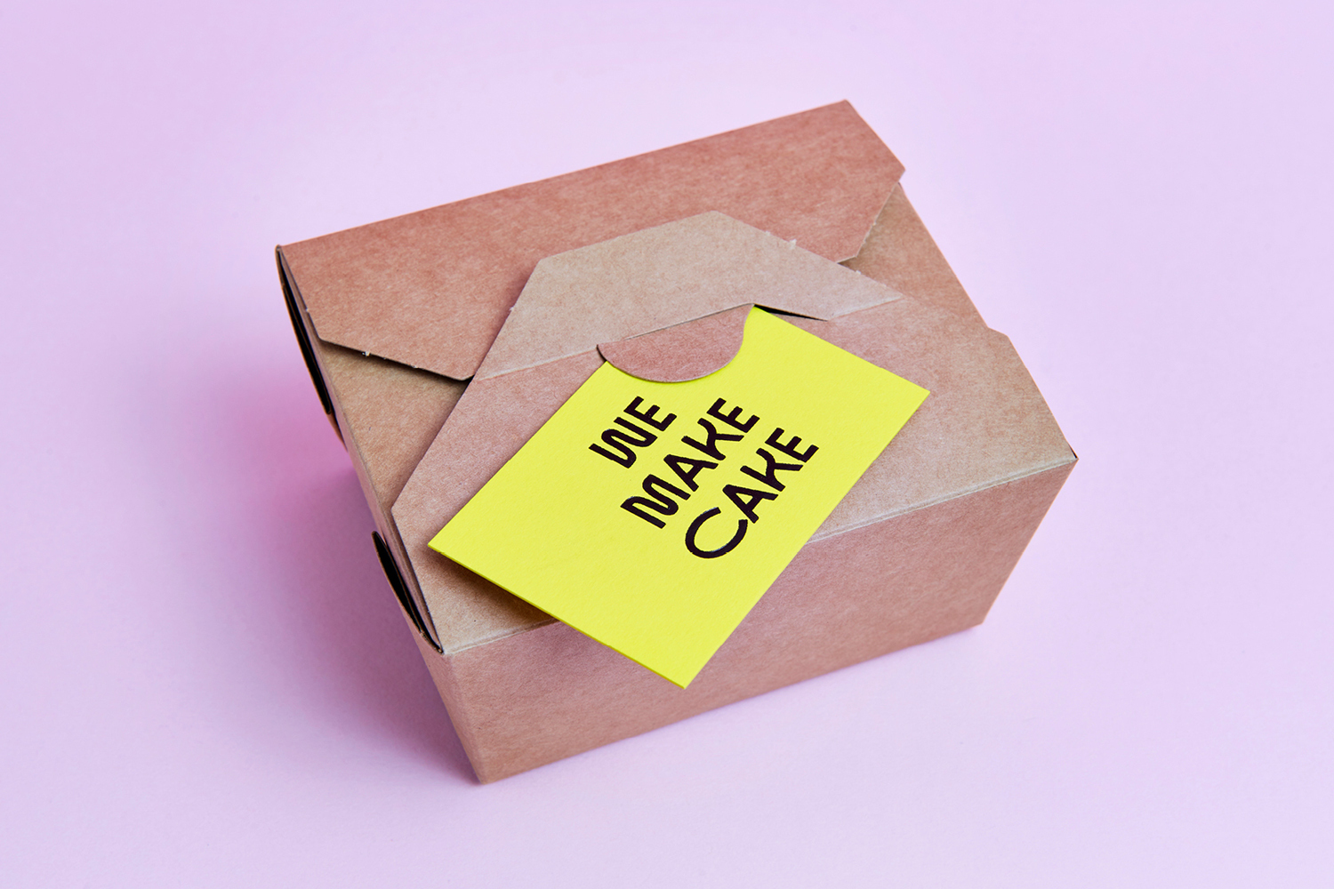 Brown cardboard cake box with a yellow label reading We Make Cake set against a pale pink background