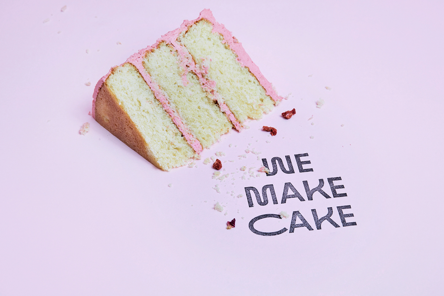Slice of layer cake with pink frosting and crumbs on a pale pink surface with branded black typography.