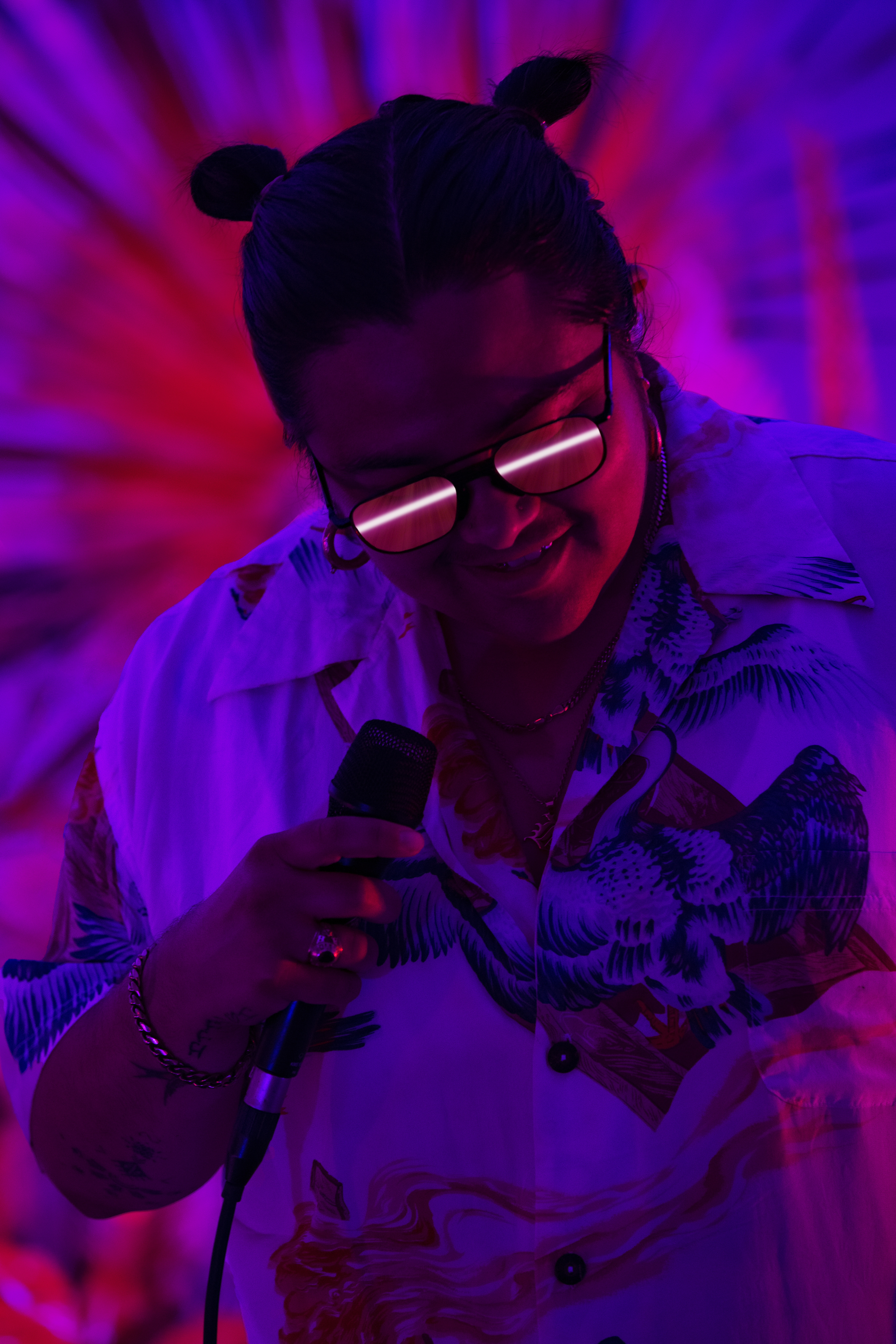 Woman singing with a microphone in a purple and red lit space. She is wearing a patterned shirt and glasses with reflected light.