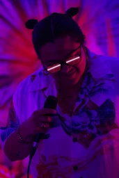 Person wearing sunglasses and patterned shirt holding a microphone under vibrant purple and red lighting