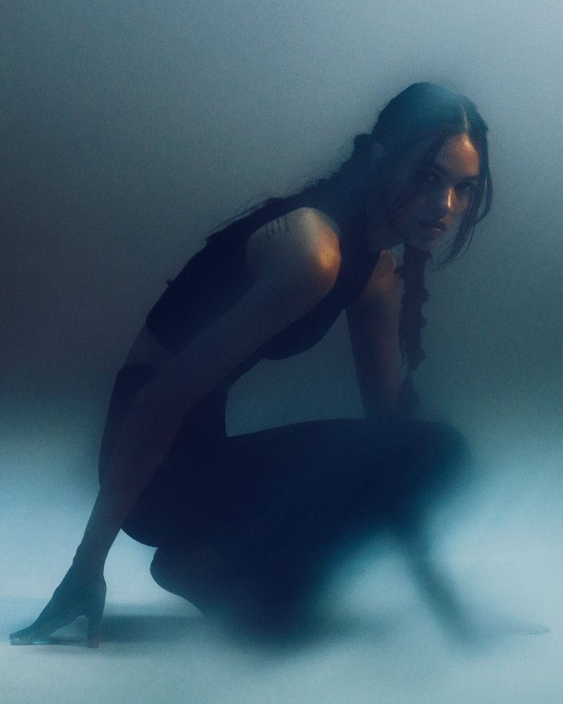 Woman crouching in black clothing with braided hair, captured in a dimly lit studio with a hazy atmosphere, exhibiting a sporty aesthetic.