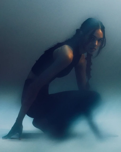 Woman crouching in black clothing with braided hair, captured in a dimly lit studio with a hazy atmosphere, exhibiting a sporty aesthetic.