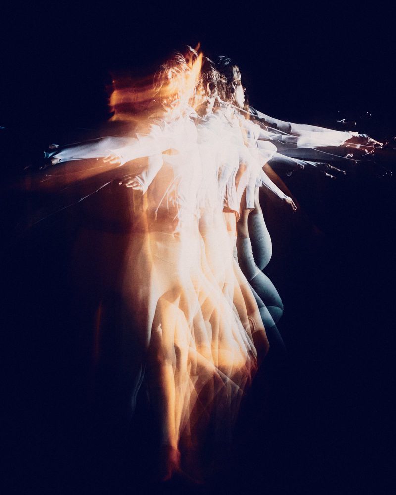 Multiple exposure photo of a woman with motion blur effect, dressed in white in a dark indoor setting, creating an ethereal, dynamic image.