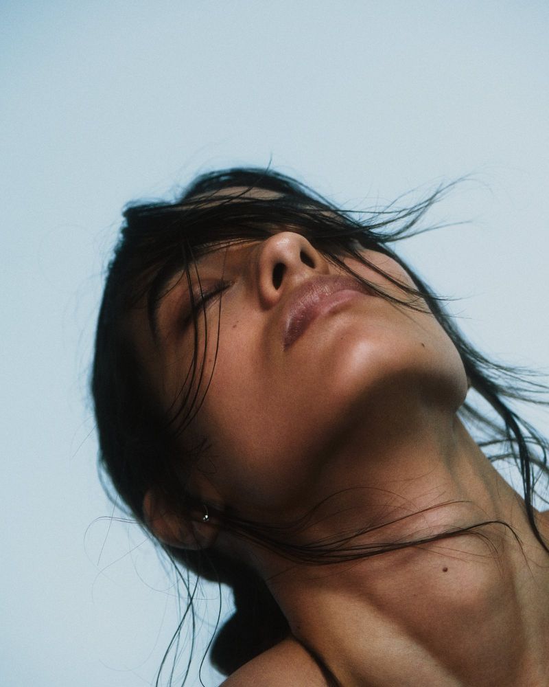 Adult female with dark hair tilted back against a pale sky. Eyes closed, neutral facial expression, medium close-up shot, natural lighting.