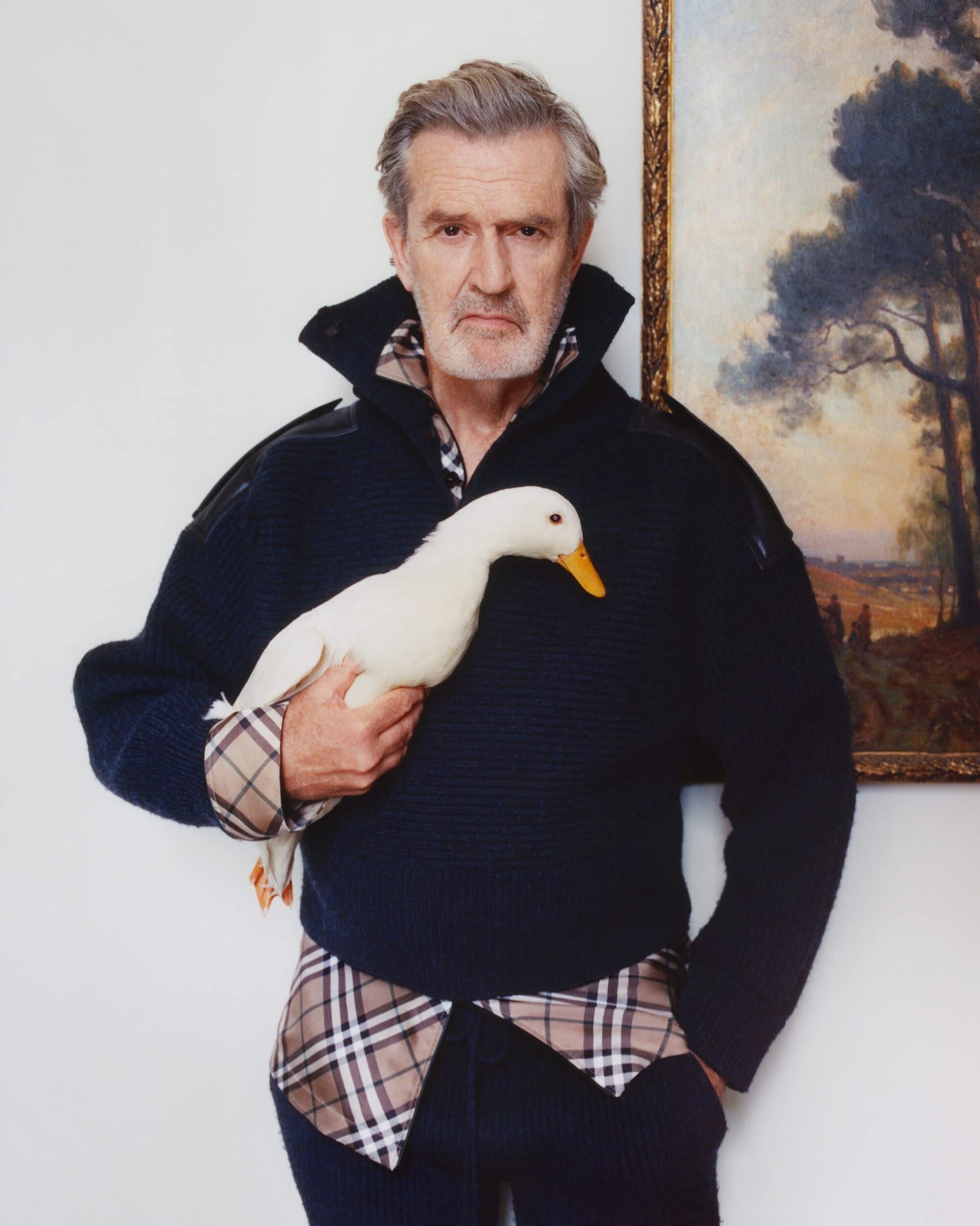 An older man wearing a dark sweater and plaid shirt holding a white duck against a light wall next to a framed landscape painting