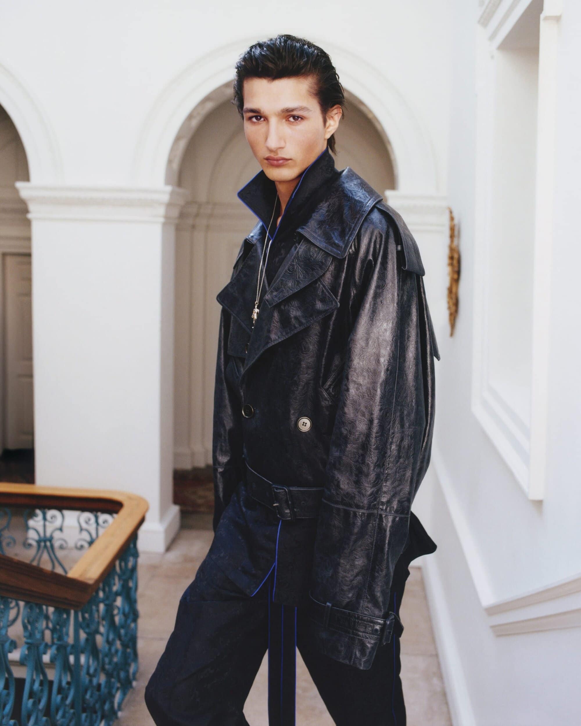 A man wearing a dark glossy leather trench coat standing on a staircase inside a grand hall with architectural arched walls.