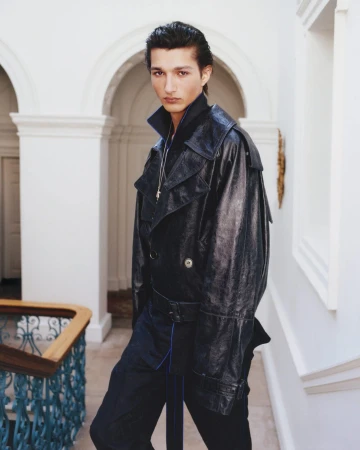 A man wearing a dark glossy leather trench coat standing on a staircase inside a grand hall with architectural arched walls.