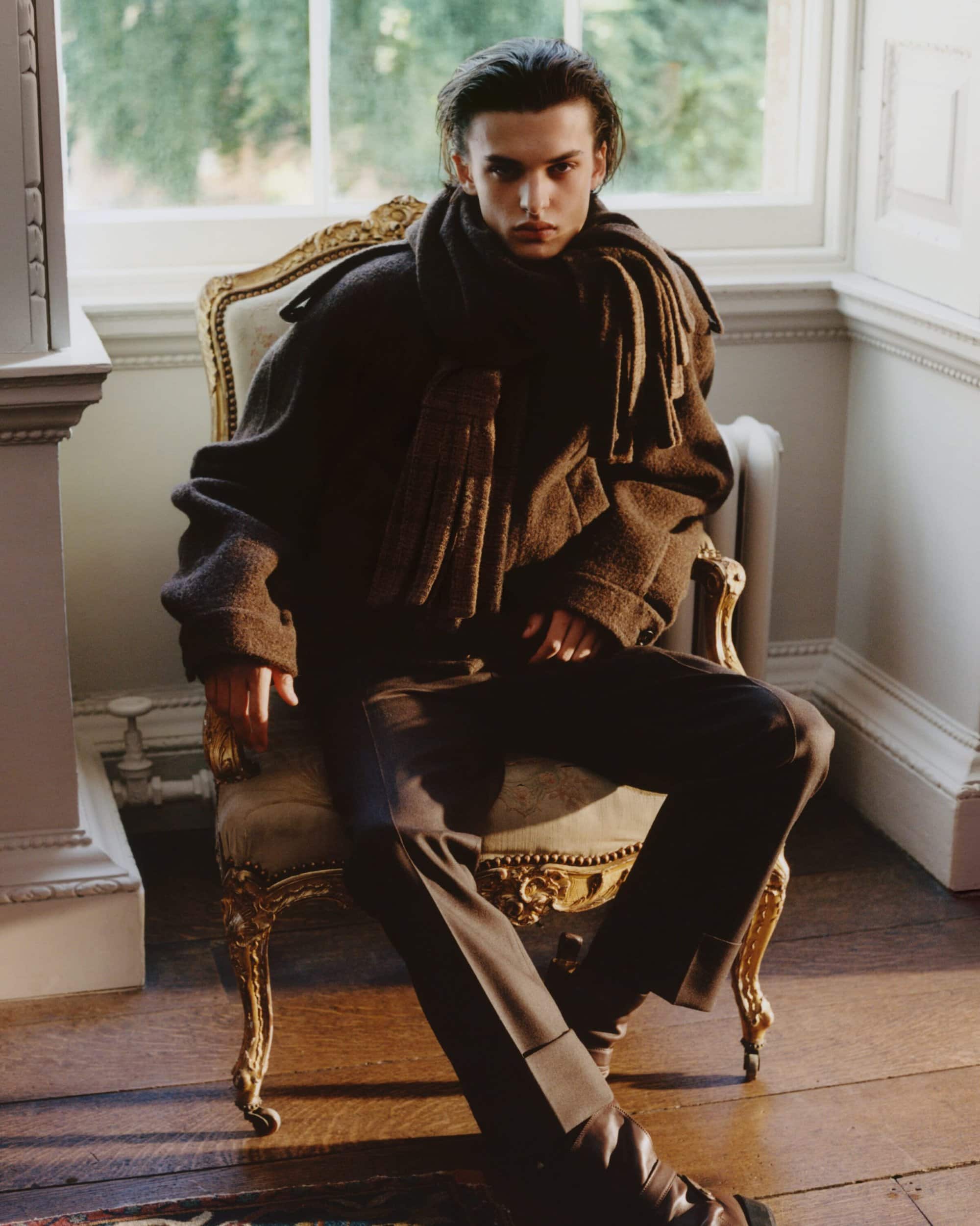A man wearing a heavy dark wool coat and scarf, seated in an ornate armchair inside a classic room with large windows.