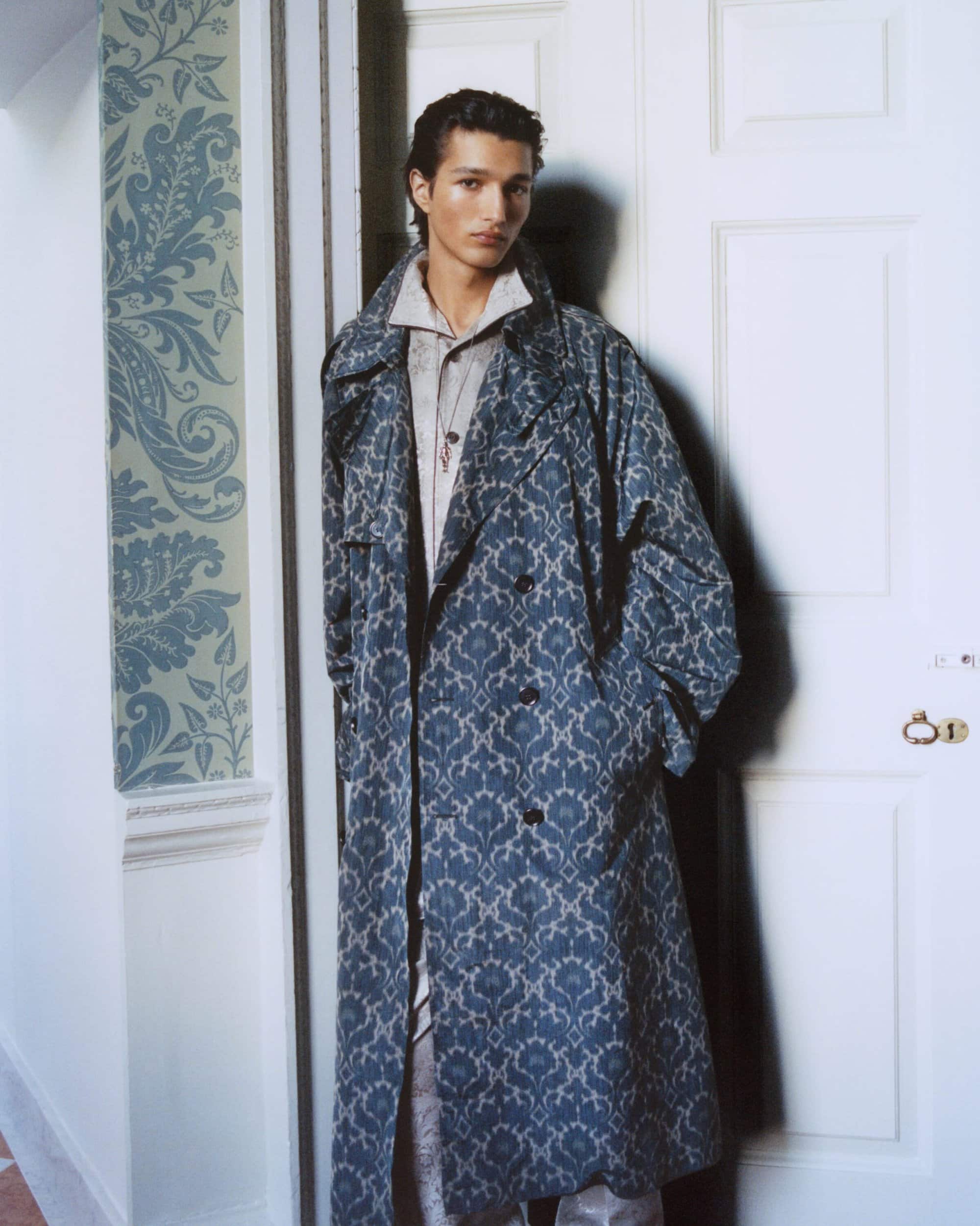 Man wearing a blue patterned long coat leaning against a white doorframe with floral wallpaper