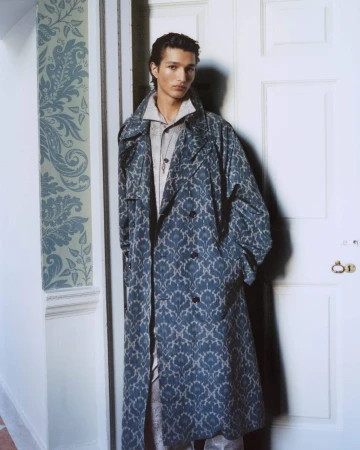 Man wearing a blue patterned long coat leaning against a white doorframe with floral wallpaper