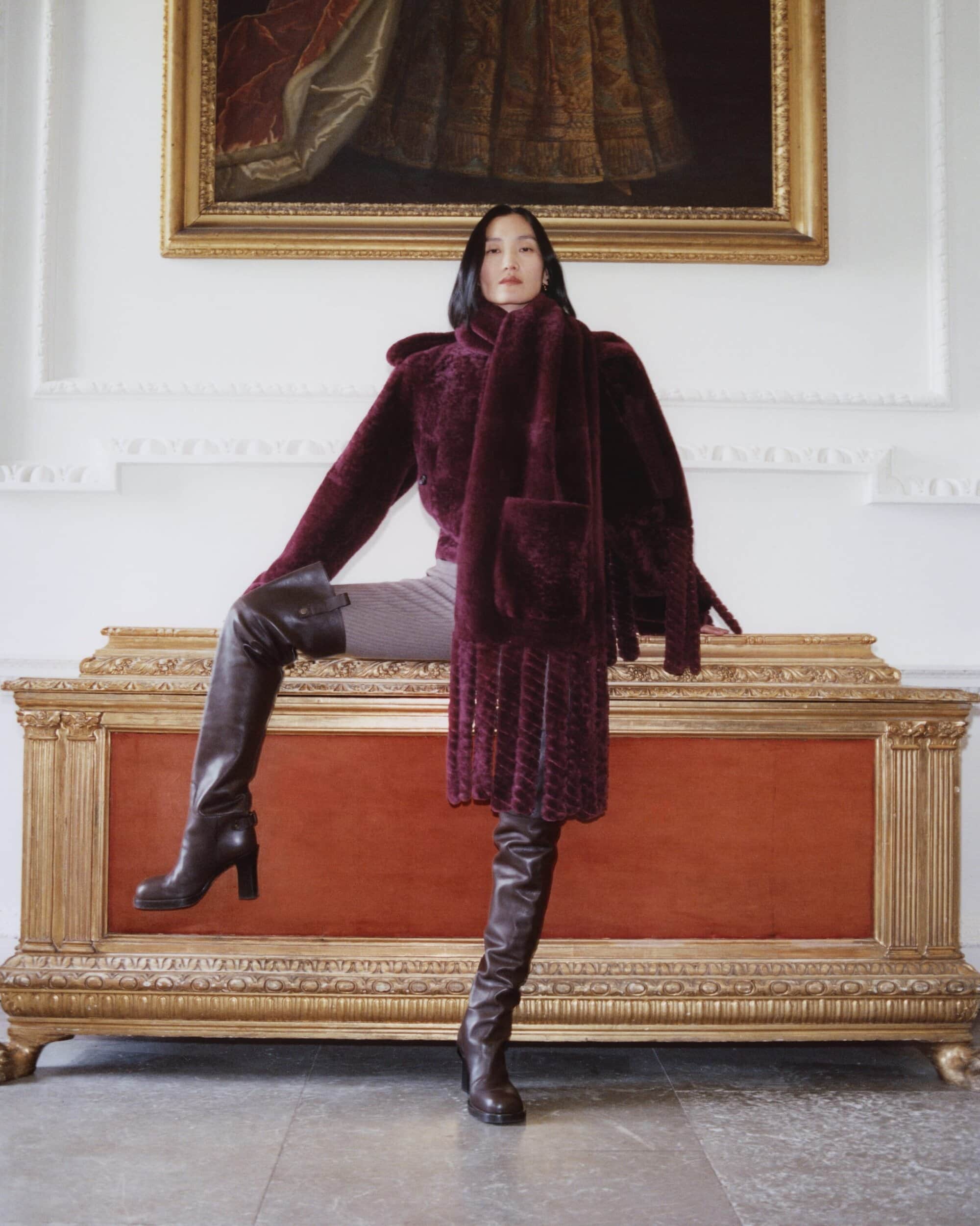 Woman wearing a burgundy textured coat and tall leather boots standing on a large wooden and red chest interior