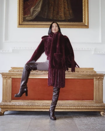 Woman wearing a burgundy textured coat and tall leather boots standing on a large wooden and red chest interior