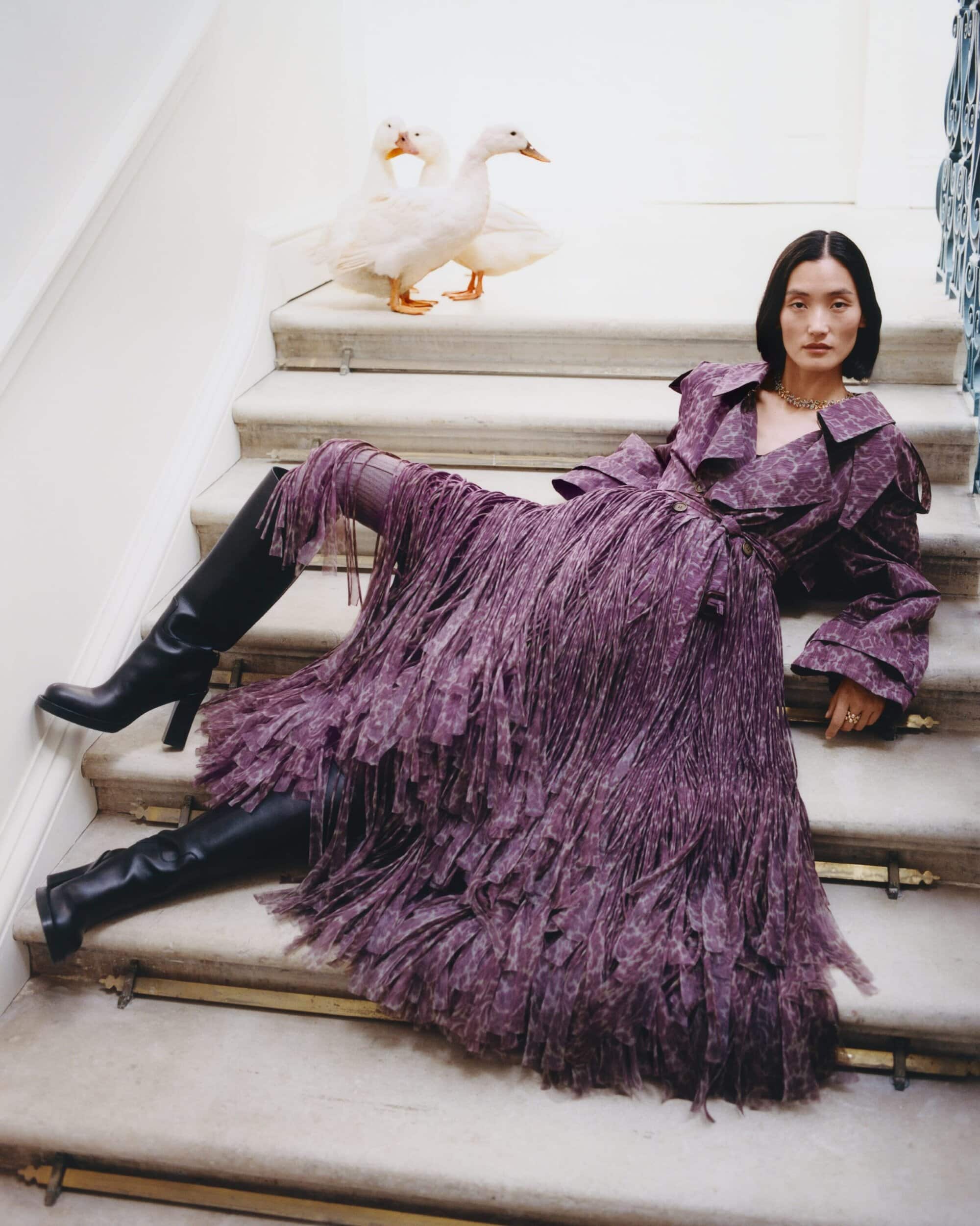 Model wearing purple fringed coat and black boots reclining on marble stairs with white ducks behind