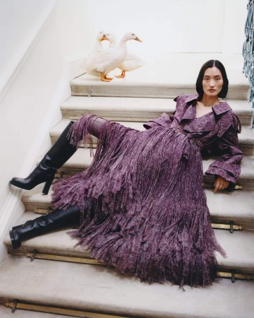 Model wearing purple fringed coat and black boots reclining on marble stairs with white ducks behind