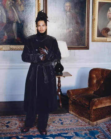 Man wearing a long dark coat standing in an interior room with framed portraits and a brown armchair