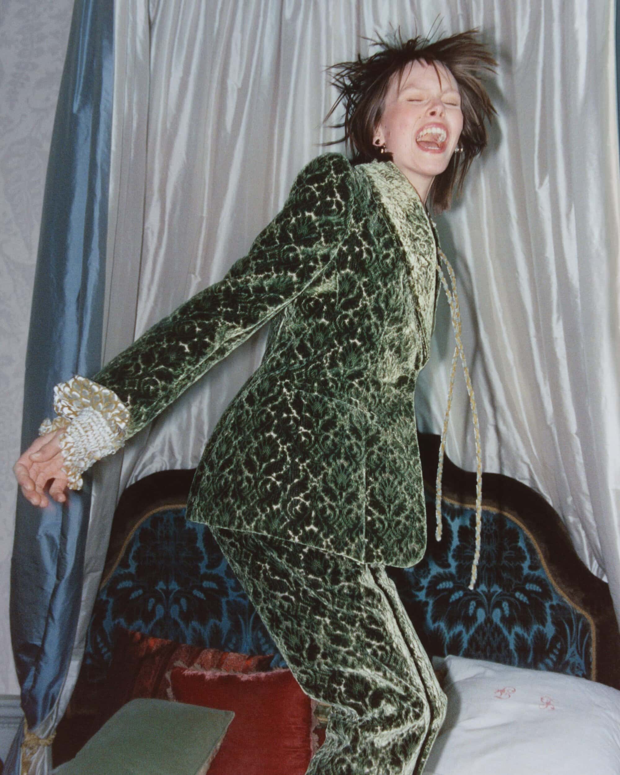 Model laughing mid-jump wearing a patterned green velvet suit indoors against draped curtains.