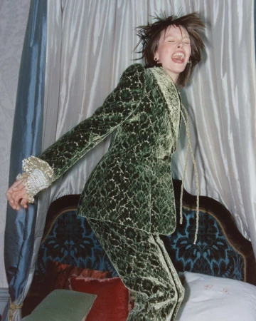 Model laughing mid-jump wearing a patterned green velvet suit indoors against draped curtains.