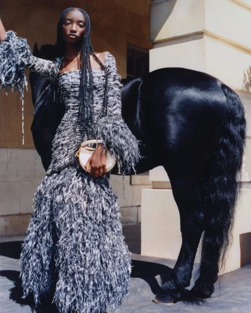 Model with long braided hair wearing a feathered grey dress standing beside a black horse