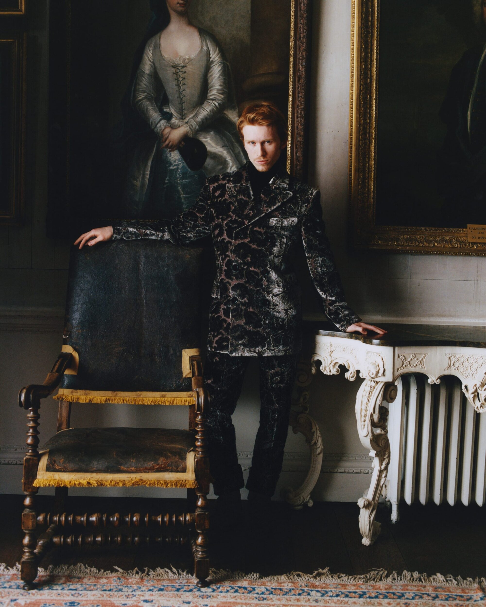 Man with red hair wearing a patterned blazer standing in an interior with historic framed paintings and antique furniture.