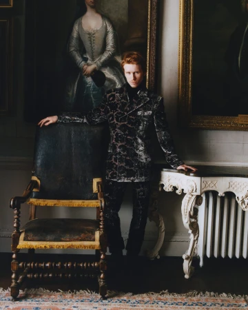 Man with red hair wearing a patterned blazer standing in an interior with historic framed paintings and antique furniture.