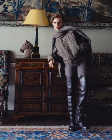 Model leaning against a wooden cabinet in a grey jacket, matching scarf, and tall dark leather boots