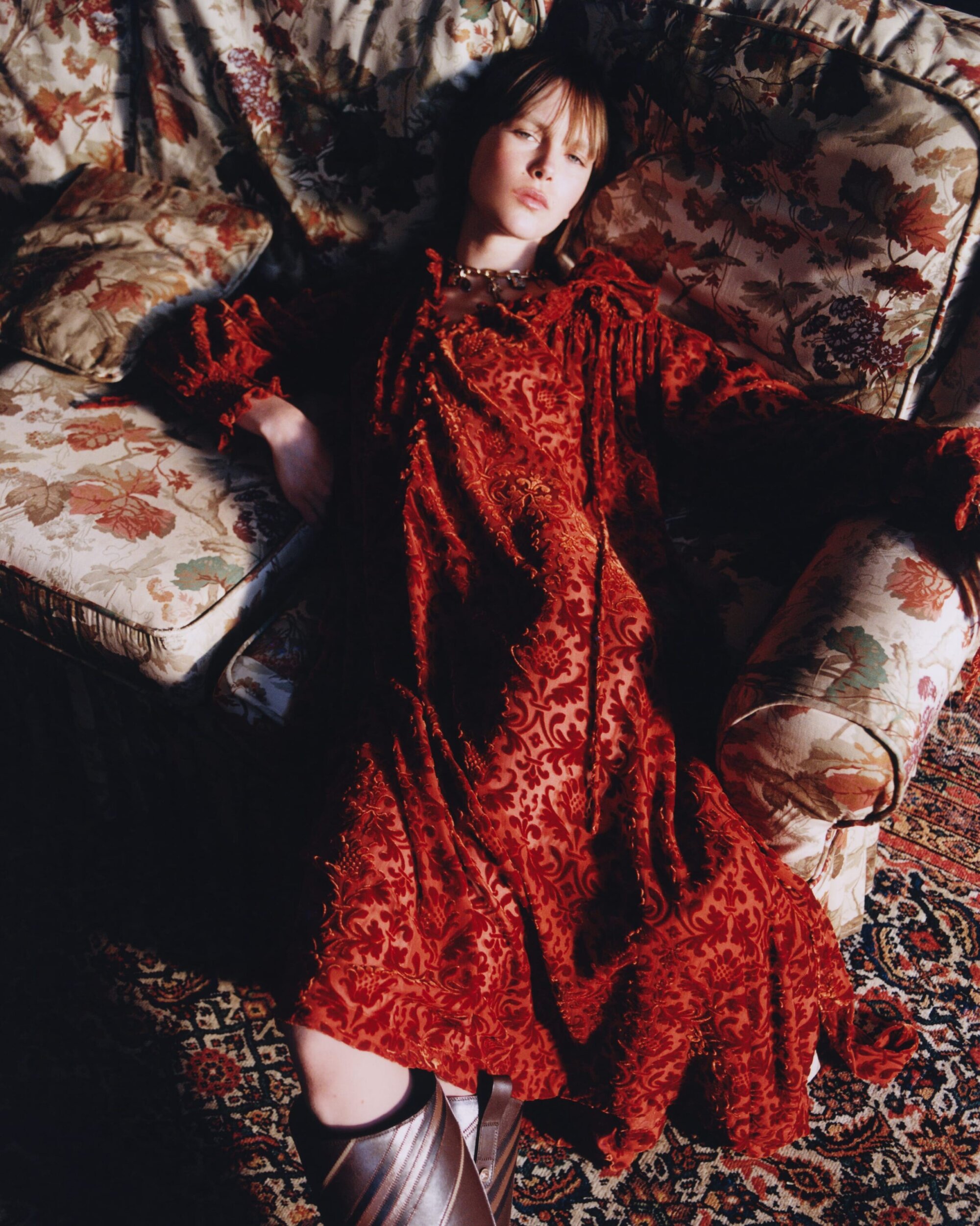 Model wearing a patterned red dress reclined on a floral upholstered sofa