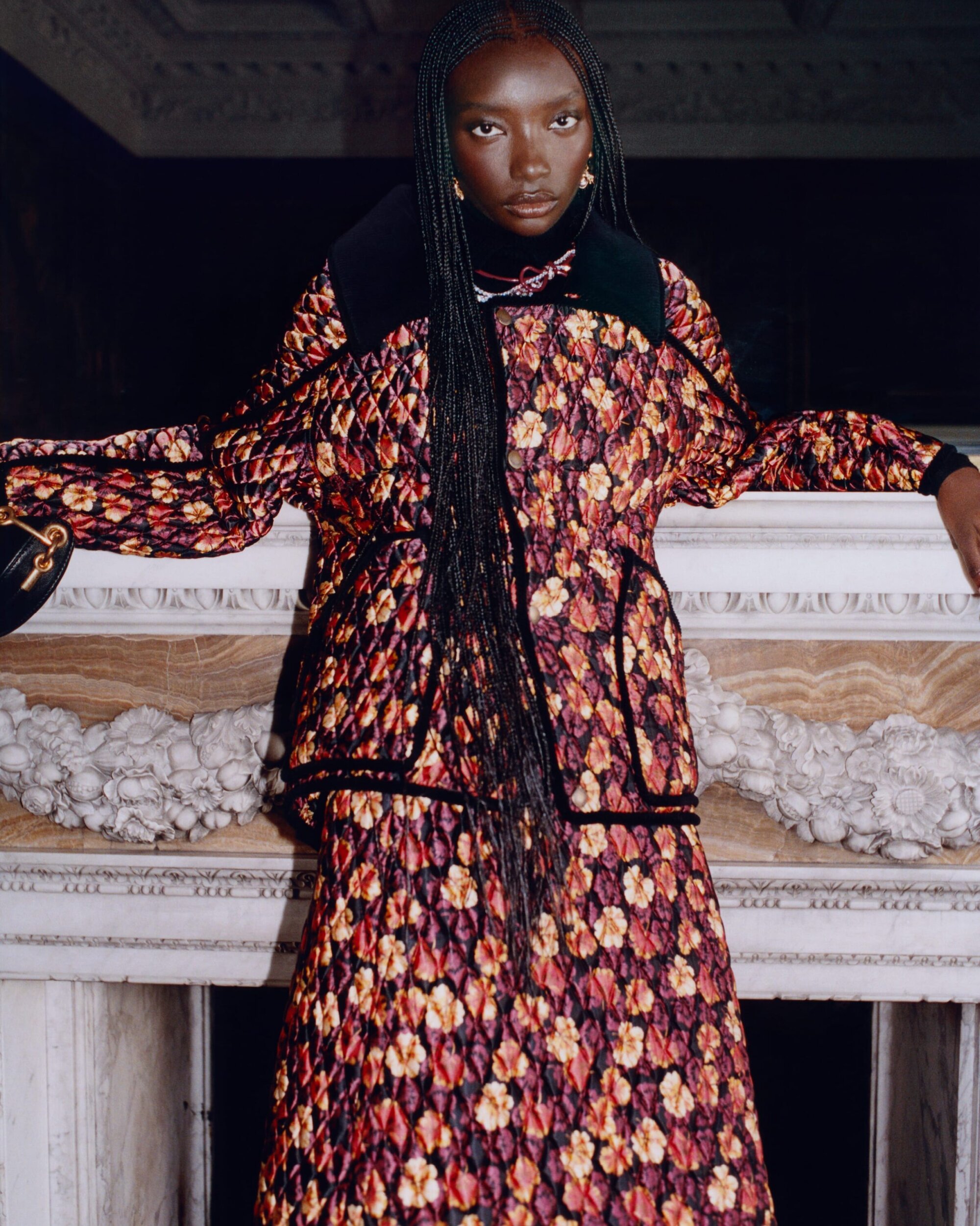Model with long braided hair wearing a quilted floral jacket leaning against a carved stone mantelpiece