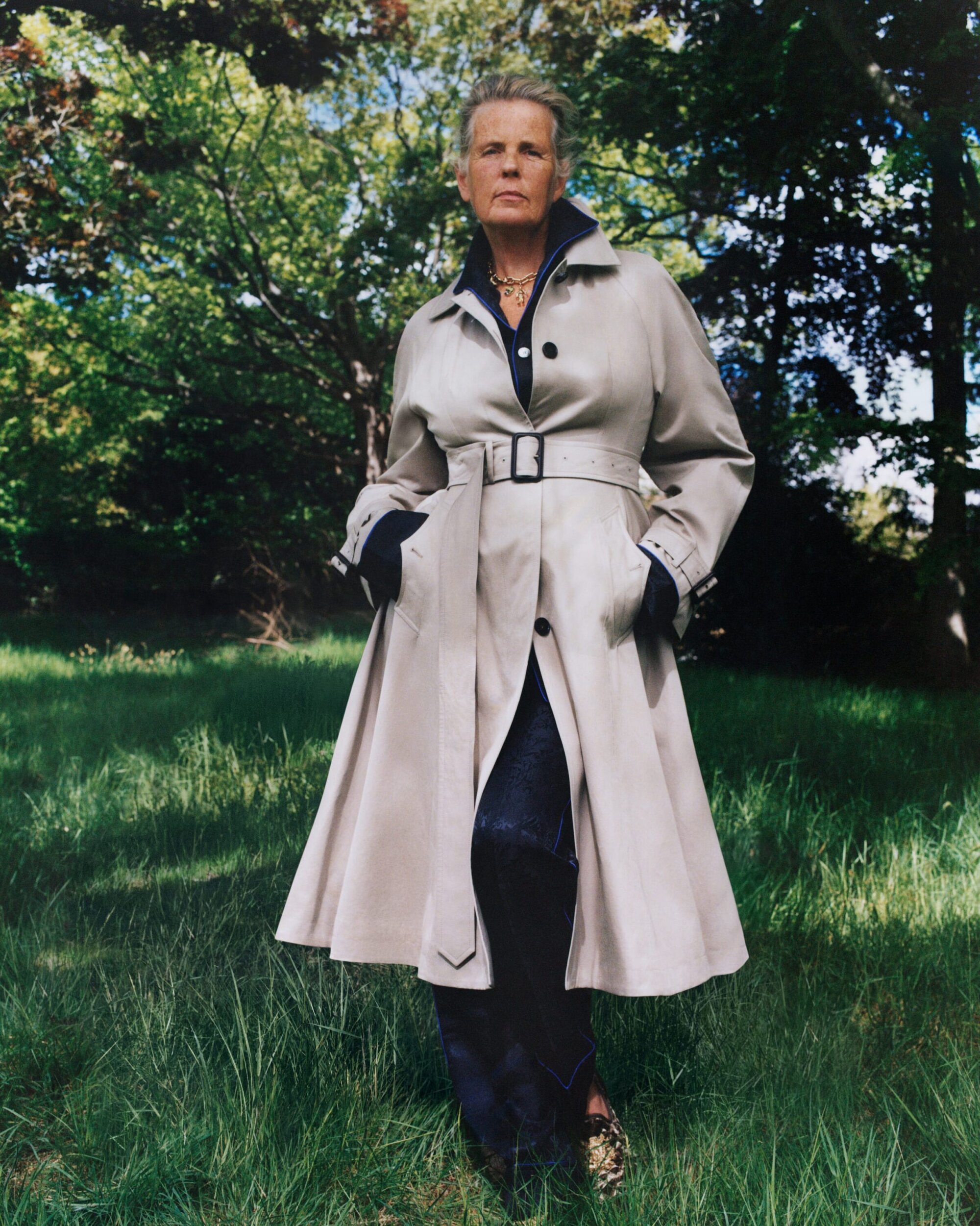 Model standing outdoors in a park wearing a beige belted trench coat over dark clothing, shot by Sam Rock for Burberry.