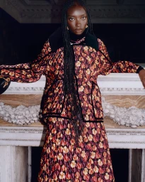 Model with long braided hair wearing a quilted floral jacket leaning against a carved stone mantelpiece
