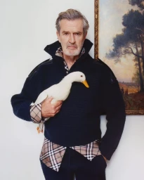 An older man wearing a dark sweater and plaid shirt holding a white duck against a light wall next to a framed landscape painting