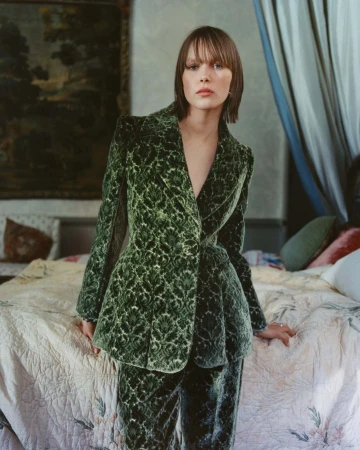 Model standing by a bed wearing a green patterned velvet blazer and matching trousers in a furnished room