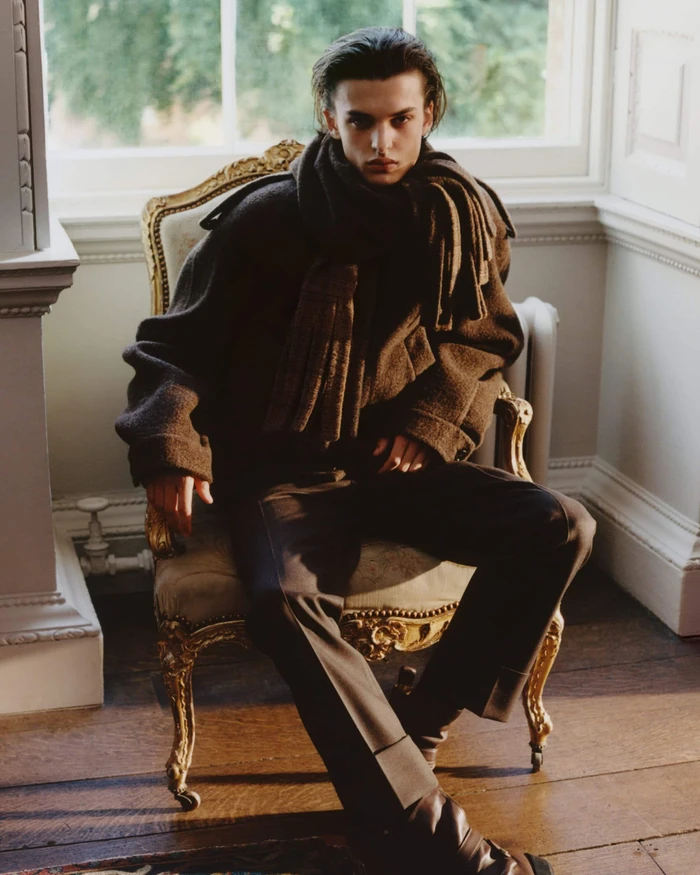 A man wearing a heavy dark wool coat and scarf, seated in an ornate armchair inside a classic room with large windows.