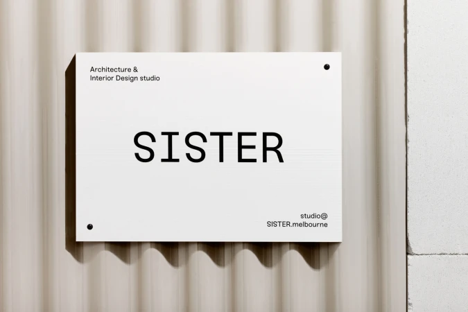 White rectangular sign with black text reading SISTER mounted on a fluted beige wall surface.