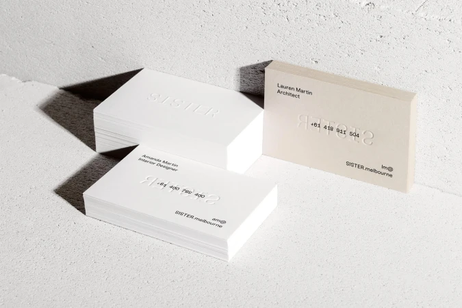 Stacks of minimalist white business cards with embossed text on a textured concrete surface under hard studio lighting.