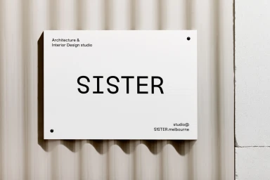 White rectangular sign with black text reading SISTER mounted on a fluted beige wall surface.