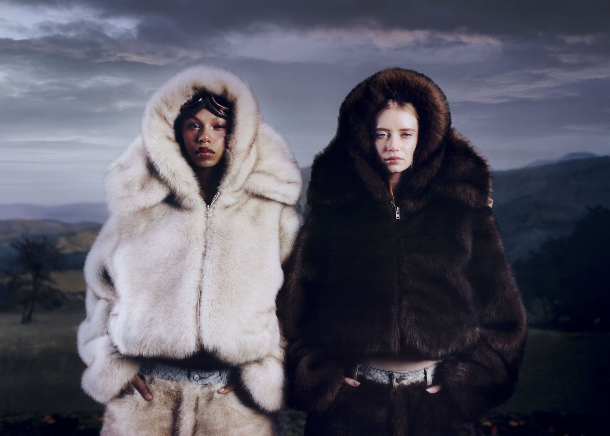 Two models wearing plush hooded winter coats posing outdoors against a dark mountainous landscape under an overcast sky.