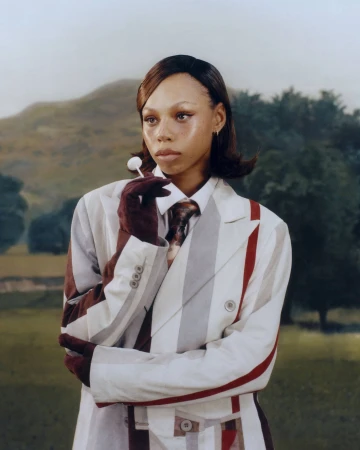 Model wearing a striped suit and dark gloves holding a lollipop against an outdoor landscape background for KidSuper Fall 2025.