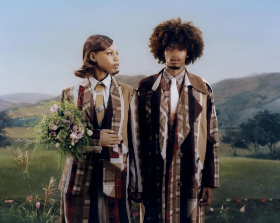 Two models wearing plaid patterned outerwear standing together in a grassy landscape holding a floral bouquet