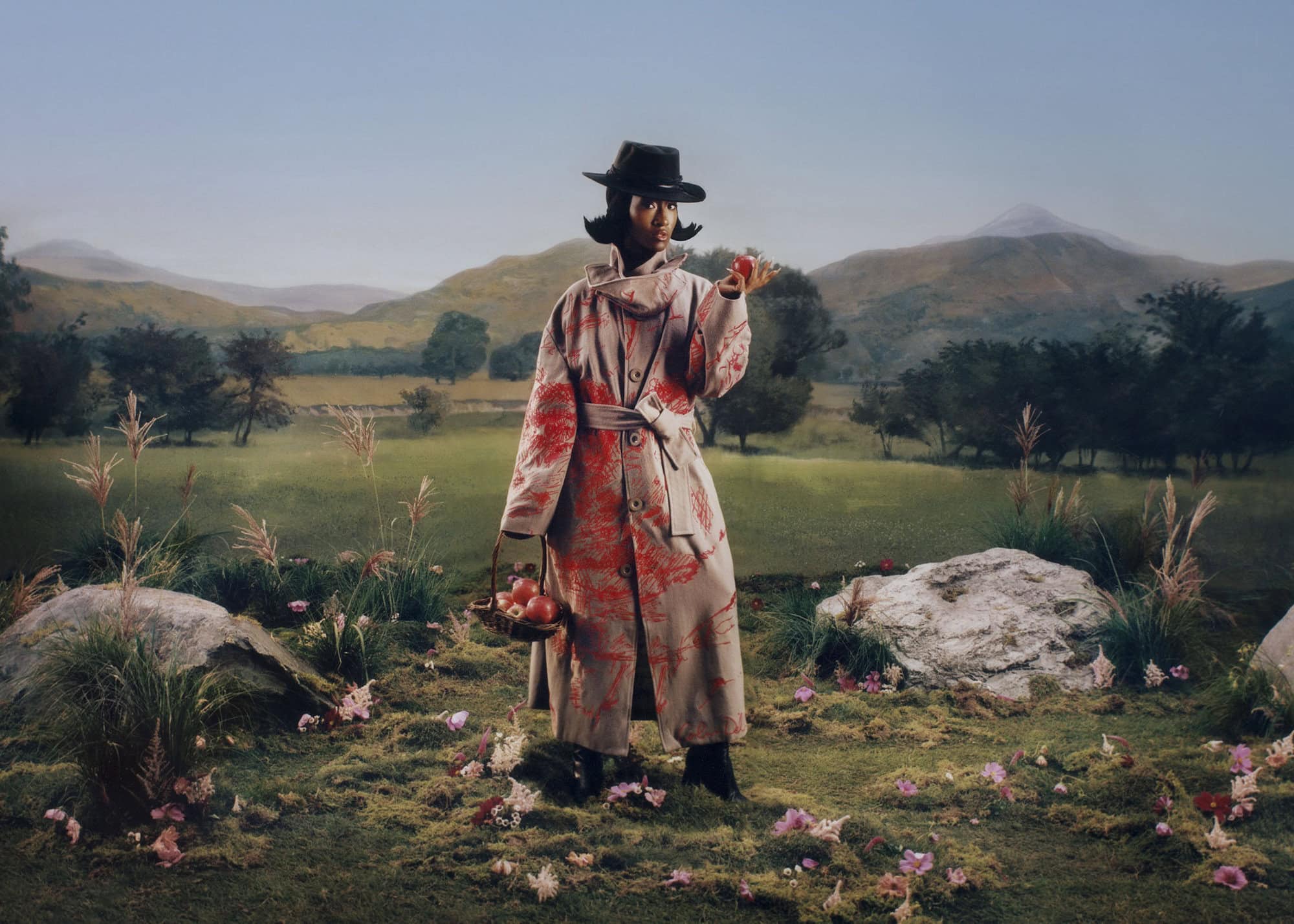 Model in patterned trench coat and hat holding a red apple standing in a painted landscape by KidSuper
