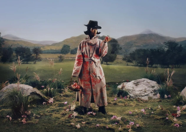 Model in patterned trench coat and hat holding a red apple standing in a painted landscape by KidSuper