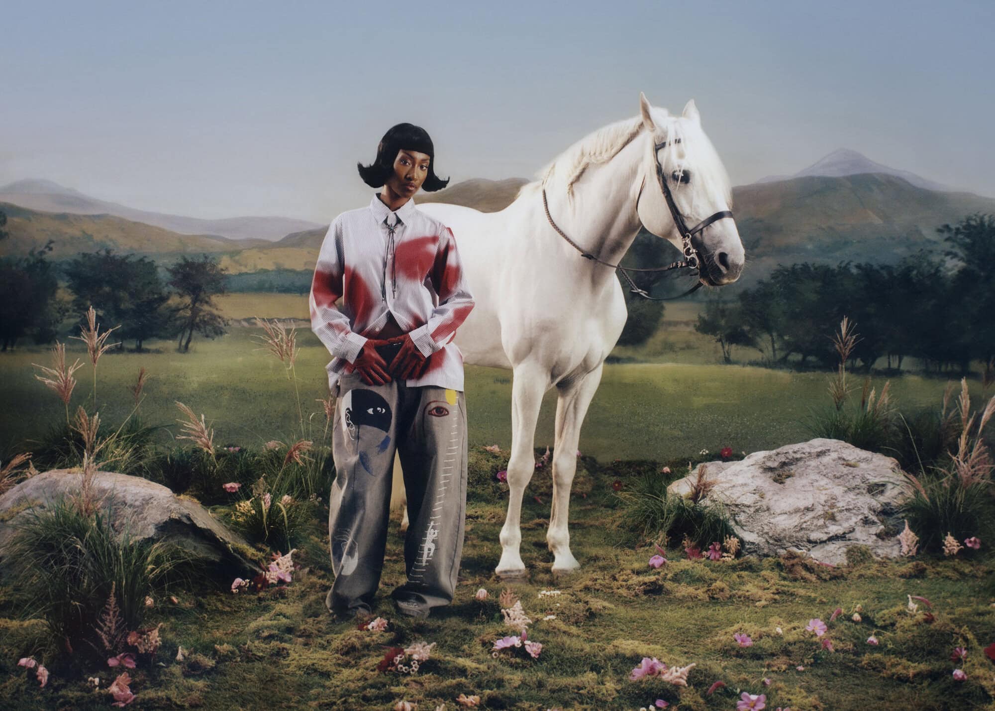 Model standing in a painted landscape next to a white horse wearing graphic trousers and shirt for KidSuper campaign