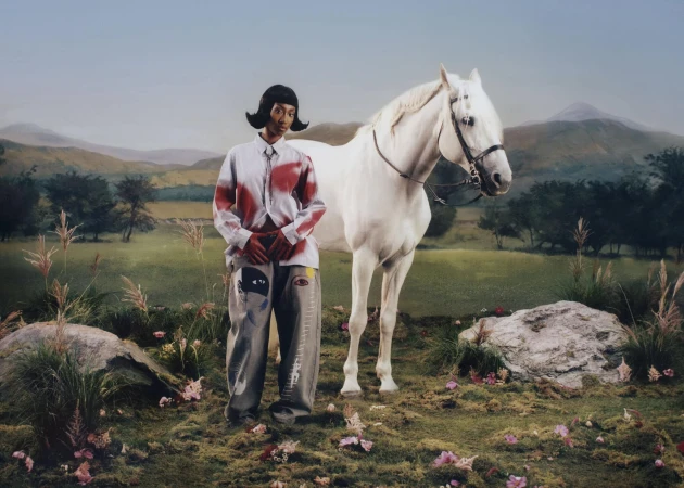Model standing in a painted landscape next to a white horse wearing graphic trousers and shirt for KidSuper campaign