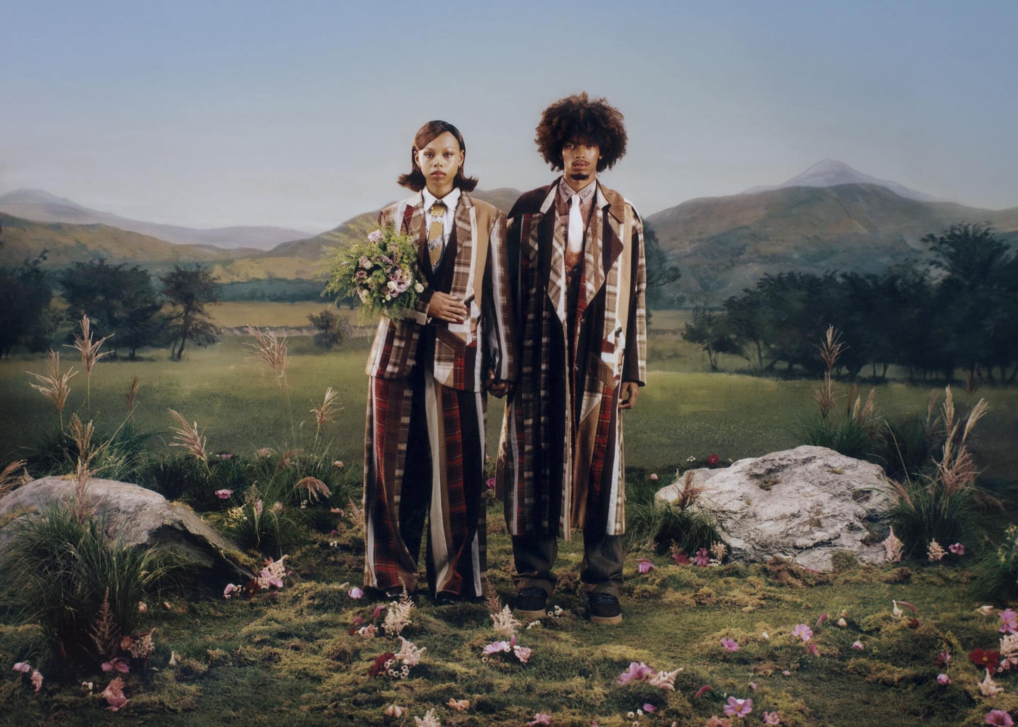 Two models wearing patterned plaid suits and coats standing in a meadow with mountains in the background