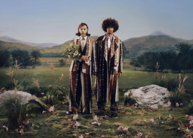 Two models wearing patterned plaid suits and coats standing in a meadow with mountains in the background