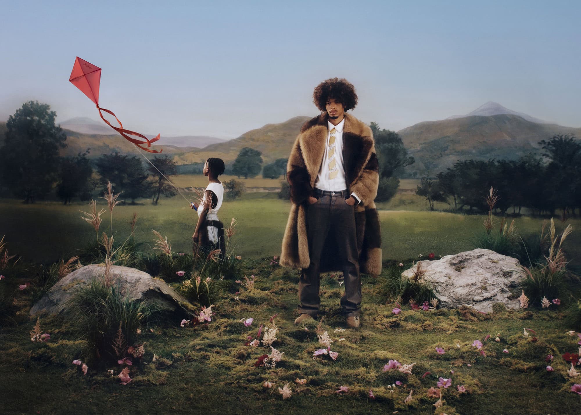 Man in fur coat standing in a grassy field with mountains in the background and a person flying a kite